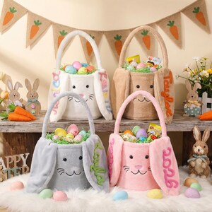 Puede incluir: Cuatro cestas de Pascua con forma de conejito en blanco, beige, gris y rosa, llenas de huevos y golosinas de colores. Las cestas tienen orejas largas y caras sonrientes. Una pancarta con diseños de zanahorias cuelga encima.