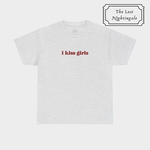 May include: A light gray short-sleeved t-shirt featuring the text i kiss girls in a dark red serif font centered on the chest. The shirt has a crew neckline and a standard fit. A small rectangular logo with The Lost Nightingale text is visible.