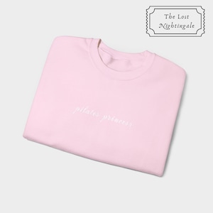 May include: A light pink sweatshirt with the embroidered text "pilates princess" in white script. The sweatshirt is folded neatly and has a crew neck. The upper right corner features a black and white logo that reads "The Lost Nightingale."