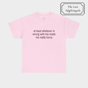 May include: A light pink short-sleeved t-shirt featuring the dark text "at least whatever is wrong with me made me really funny" printed on the chest. The shirt has a crew neckline and a standard fit, with a small brand label inside the collar.