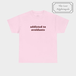 Puede incluir: Una camiseta de manga corta de color rosa claro con el texto en rojo oscuro "addicted to avoidants" impreso en el centro del pecho. La camiseta incluye una pequeña etiqueta interior en el cuello con el texto "The Lost Nightingale" y un logo.