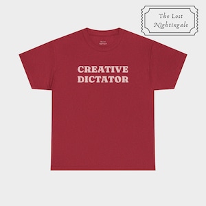 May include: A dark red short-sleeve t-shirt featuring the text CREATIVE DICTATOR in a light pink serif font across the chest. The inner neck label displays the text The Lost Nightingale, which also appears in a decorative frame above the shirt.