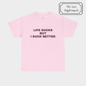 May include: A light pink short-sleeved t-shirt featuring the black text LIFE SUCKS BUT I SUCK BETTER printed on the chest. The shirt has a crew neckline and a standard fit. A small logo box in the top right corner reads The Lost Nightingale.