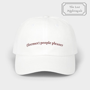 Può includere: Un cappellino da baseball bianco con visiera curva. Il cappellino presenta la frase "(former) people pleaser" ricamata in rosso scuro. Nell'angolo in alto a destra è presente un logo in bianco e nero con la scritta "The Lost Nightingale".