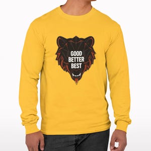 Può includere: Maglietta a maniche lunghe giallo senape con grafica a testa di orso nera e rossa e la scritta "GOOD BETTER BEST". La grafica dell'orso ha denti affilati e un design dettagliato. La maglietta è realizzata in materiale morbido.