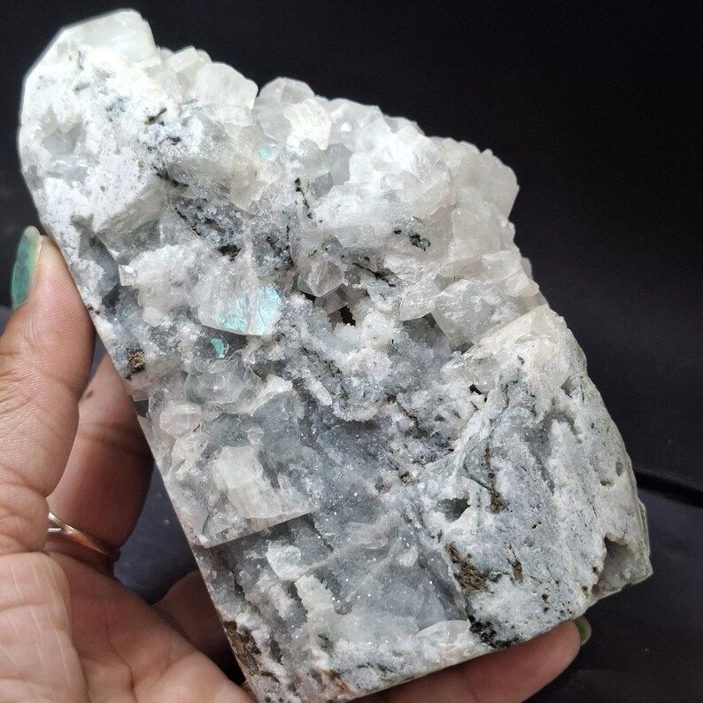 May include: A large, raw mineral specimen with a mix of white, gray, and black tones. The surface has a crystalline structure with small, light blue inclusions. The stone is held in a hand, showcasing its natural texture and color variations.