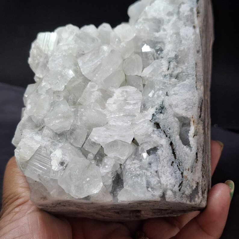 May include: A large, raw crystal cluster with a light gray and white color palette. The crystal formation features numerous small, faceted crystals clustered together, with a rough, textured exterior. The overall shape is rectangular, with a dark gray edge.
