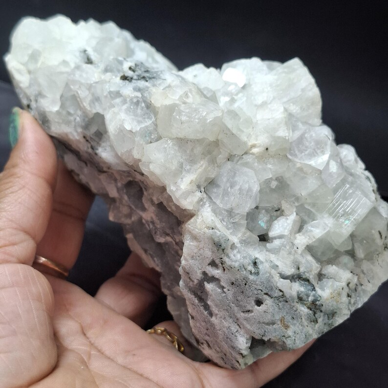 May include: A large, raw, crystalline mineral specimen. The stone is primarily white with areas of gray and black. The surface has a rough, textured appearance with visible crystal formations. The mineral is held in a hand.