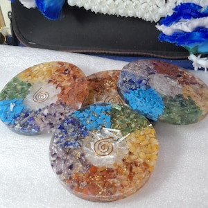 May include: Four colorful, oval-shaped orgone energy generators. Each features a spiral design at the center, surrounded by a mosaic of gemstones in various colors, including blue, green, orange, and purple. The stones are embedded in a clear resin.