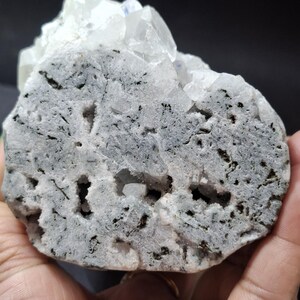 May include: A close-up of a raw, uncut mineral specimen. The stone has a light gray base with dark gray and black inclusions and several small holes. Clear, crystalline formations are visible on top.
