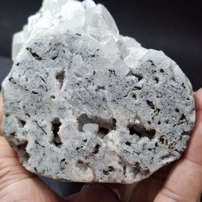 May include: A close-up of a raw, uncut mineral specimen. The stone has a light gray base with dark gray and black inclusions and several small holes. Clear, crystalline formations are visible on top.