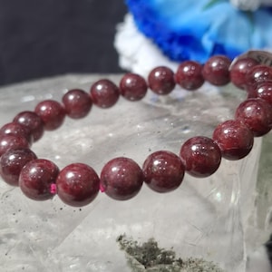 Peut inclure: Un bracelet de perles bordeaux. Il est composé de perles rondes et polies, enfilées sur un cordon élastique rose. Les perles sont d'un rouge-brun profond. Le bracelet est présenté sur une surface claire et texturée.