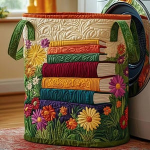May include: Quilted laundry basket with a floral and book design. The basket features a stack of colorful books and embroidered flowers in shades of yellow, pink, and orange. Green handles and trim complete the design.