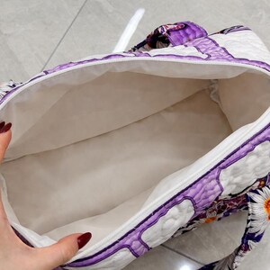 May include: A quilted duffel bag featuring a vibrant purple and white pattern with butterfly and daisy motifs. The bag has a white interior lining, a zippered top closure, and matching patterned fabric handles attached to the exterior sides.