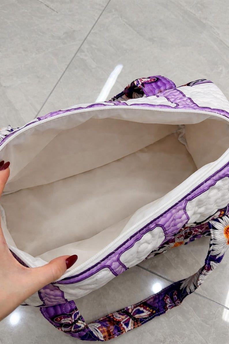May include: A quilted duffel bag featuring a vibrant purple and white pattern with butterfly and daisy motifs. The bag has a white interior lining, a zippered top closure, and matching patterned fabric handles attached to the exterior sides.