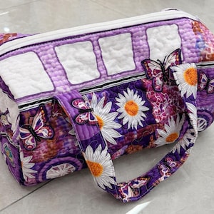 May include: A quilted barrel-shaped bag featuring a purple and white color scheme with a pattern of daisies and butterflies. The bag has a white zipper closure along the top, two fabric handles, and a series of white rectangular quilted panels.