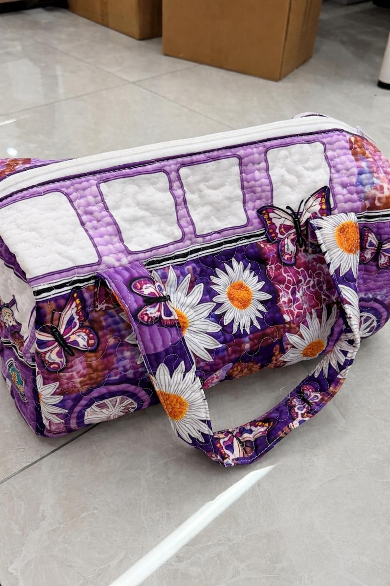 May include: A quilted barrel-shaped bag featuring a purple and white color scheme with a pattern of daisies and butterflies. The bag has a white zipper closure along the top, two fabric handles, and a series of white rectangular quilted panels.