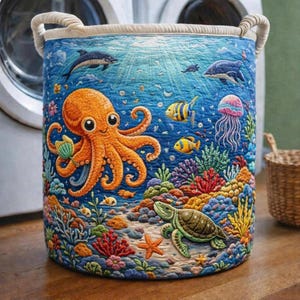 May include: A cylindrical laundry hamper with a vibrant underwater scene. The design features an orange octopus, dolphins, colorful fish, a sea turtle, and coral reefs against a blue background. The hamper has rope handles and a cream-colored rim.