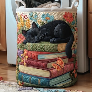 May include: A decorative laundry hamper featuring a black cat sleeping on a stack of colorful books. The hamper is adorned with floral embroidery in shades of yellow, orange, and blue, with a butterfly accent. The hamper has a cream-colored interior and handles.