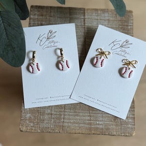 May include: Two pairs of white baseball-shaped earrings with red stitching details. One pair features gold-tone hoop findings, while the other pair includes gold-tone bow accents. Each pair is displayed on a white card labeled with KC Ketty's Creations.