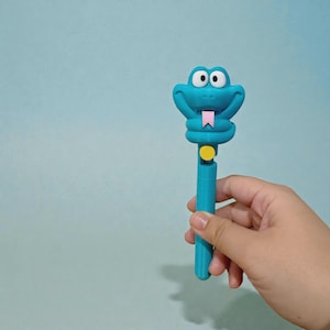 May include: A turquoise pen with a cartoon snake head design. The snake has large eyes, a pink tongue, and a yellow button on the pen's body. The pen is held against a light blue background.