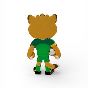 Puede incluir: Figura de un le&oacute;n de dibujos animados con un uniforme de f&uacute;tbol verde, sosteniendo un bal&oacute;n de f&uacute;tbol. El le&oacute;n tiene un cuerpo marr&oacute;n, orejas marrones y zapatos negros y amarillos. Vista trasera.