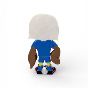 Puede incluir: Una figura de dibujos animados de un futbolista, vista desde atr&aacute;s, en azul, marr&oacute;n y amarillo. La figura tiene una cabeza blanca, una camiseta azul, brazos marrones, pantalones cortos amarillos y zapatos negros y rosas. Un bal&oacute;n de f&uacute;tbol est&aacute; a los pies.