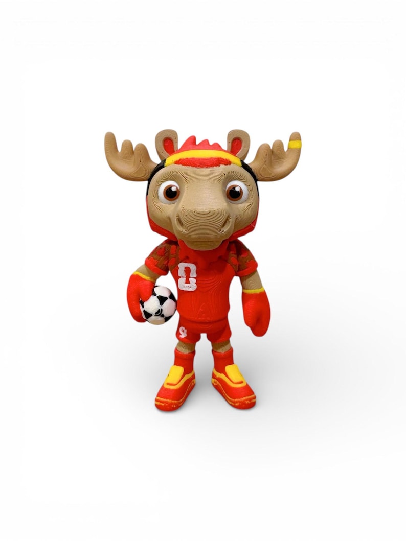 Puede incluir: Una mascota de alce de peluche con un uniforme de f&uacute;tbol rojo con el n&uacute;mero 9. La mascota tiene grandes astas marrones, una diadema negra y amarilla y sostiene un bal&oacute;n de f&uacute;tbol blanco y negro. Los zapatos son rojos y amarillos.