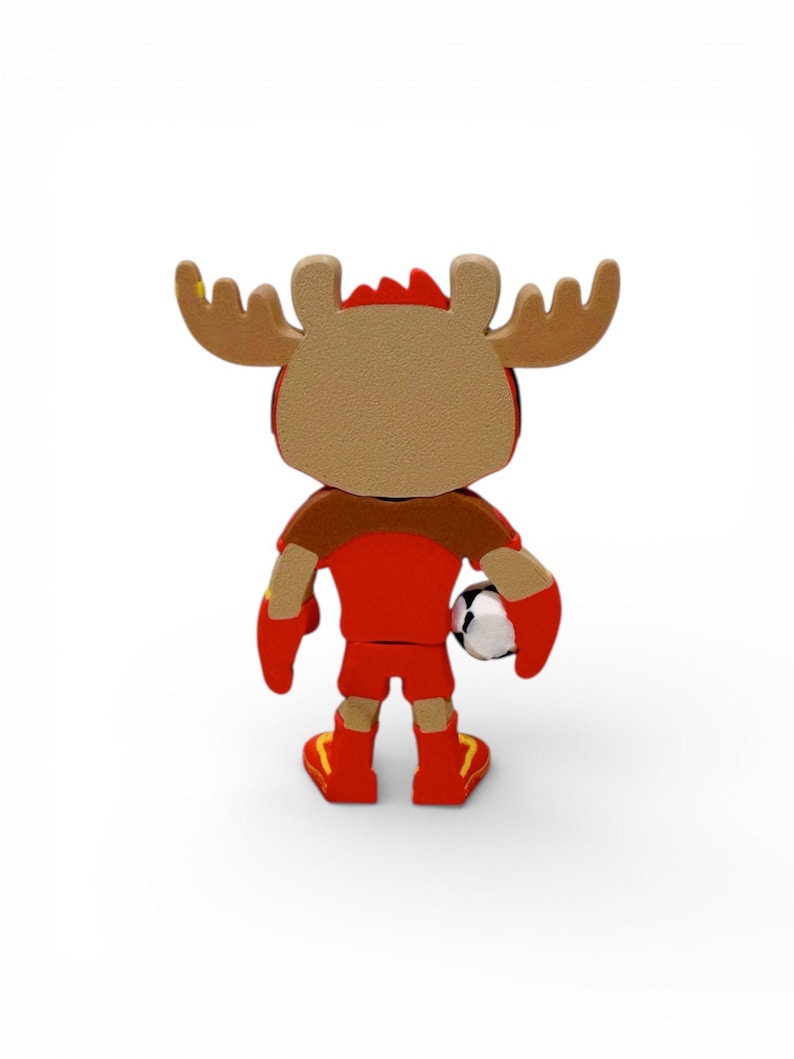 Puede incluir: Una figura de dibujos animados de un alce con uniforme de f&uacute;tbol rojo, sosteniendo un bal&oacute;n de f&uacute;tbol. El alce tiene cuernos marrones y un cuerpo rojo y marr&oacute;n. La figura est&aacute; sobre una superficie blanca.