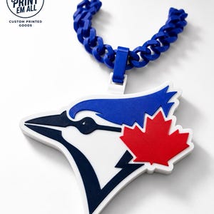 May include: A blue and white pendant necklace featuring the Toronto Blue Jays logo with a red maple leaf. The necklace has a chunky blue chain and a white clasp. The text "Gotta Print Em All" is in a circle.