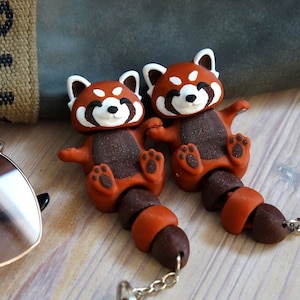 Può includere: Due portachiavi a forma di panda rosso fatti a mano. Ogni portachiavi presenta un panda rosso con viso bianco, corpo marrone scuro e una lunga coda segmentata. I portachiavi sono attaccati a una catena d'argento.