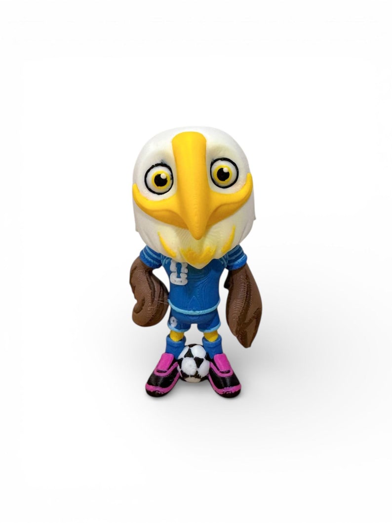 Puede incluir: Figura de un &aacute;guila con un uniforme de f&uacute;tbol azul con el n&uacute;mero 3. El &aacute;guila tiene un pico amarillo y alas marrones. Est&aacute; de pie sobre un bal&oacute;n de f&uacute;tbol y lleva zapatos rosas y negros.