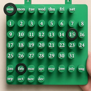 May include: A green, modular calendar with white numbers and month abbreviations. Black rings highlight the current day and month. The calendar has a grid of circular pegs for the numbers and months.