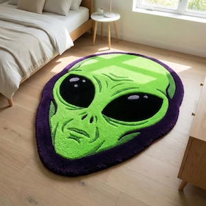 Könnte beinhalten: Ein grüner Alien-Gesichtsteppich mit großen schwarzen Augen und einem lila Rand. Der Teppich besteht aus einem weichen, plüschigen Material und ist in einer einzigartigen, auffälligen Form gestaltet. Dieser dekorative Teppich verleiht jedem Raum eine lustige, skurrile Note.