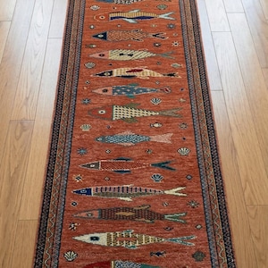Könnte beinhalten: Ein rechteckiger, terrakottafarbener Teppich mit einem sich wiederholenden Muster aus bunten Fischen. Die Fische sind in verschiedenen Designs und Farben, darunter Blau, Gelb und Rot. Der Teppich hat einen dunkelblauen Rand mit einem detaillierten Muster und Fransen.