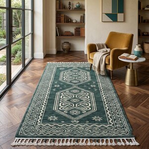 May include: A rectangular area rug in deep teal with a white geometric pattern featuring central hexagonal motifs, star shapes, and crescent moons. The rug has a decorative border and white fringe tassels along the short edges on both ends.