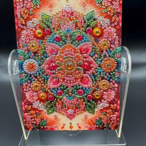 Puede incluir: Una pieza de arte rectangular decorativa con un vibrante diseño floral de mandala. La obra de arte presenta un motivo floral central rodeado de intrincados patrones en tonos de rojo, rosa, naranja y turquesa, adornado con acentos brillantes. Se exhibe en un soporte de acrílico transparente.