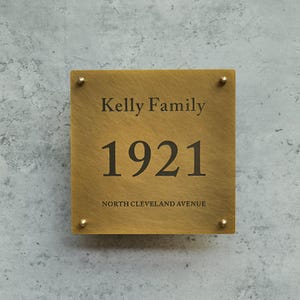 May include: A square, brass-colored house number sign with the text "Kelly Family" above the number "1921". The words "NORTH CLEVELAND AVENUE" are below the number. The sign is attached with four gold-colored screws.