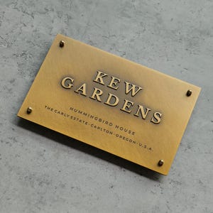 Puede incluir: Un letrero rectangular de latón con el texto en relieve "KEW GARDENS" y texto más pequeño debajo. El letrero está asegurado con cuatro tornillos de latón. El texto incluye "HUMMINGBIRD HOUSE" y "THE CARLY ESTATE - CARLTON - OREGON - U.S.A."