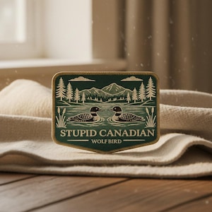 Puede incluir: Un parche verde y beige con un diseño de paisaje que presenta dos somorgujos en un lago, árboles y montañas. El texto "STUPID CANADIAN" y "WOLF BIRD" se muestra en el parche.