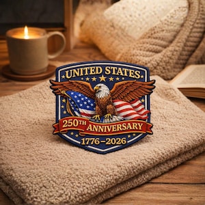 Puede incluir: Parche patriótico con un águila, la bandera estadounidense y el texto "UNITED STATES 250TH ANNIVERSARY 1776-2026". El parche es azul, rojo y dorado. La imagen está sobre una toalla beige doblada.