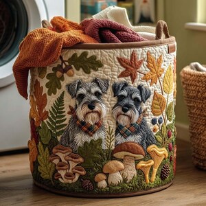 Puede incluir: Una cesta de almacenamiento redonda de tela con fondo crema acolchado, decorada con bordados de hojas de otoño, setas y dos perros Schnauzer con pañuelos a cuadros. La cesta está llena de telas plegadas de color naranja y granate.