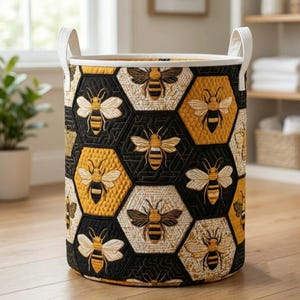 Pode incluir: Um cesto de armazenamento cilíndrico de tecido com um padrão acolchoado de favo de mel em preto, branco e amarelo. O exterior é decorado com motivos de abelhas bordados dentro de cada hexágono. Inclui duas alças verticais brancas no topo.