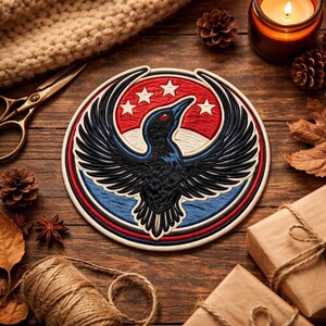 Op de afbeelding: Geborduurde patch met een gestileerde zwarte vogel met uitgespreide vleugels, rode ogen en een rood-wit-blauw cirkelvormig ontwerp. De patch bevat drie witte sterren op een rode achtergrond. Op een houten oppervlak.