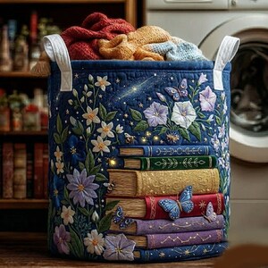 Op de afbeelding: Een cilindervormige wasmand van donkerblauwe stof met gewatteerd borduurwerk van gestapelde boeken, kleurrijke wilde bloemen en vlinders. De mand heeft twee witte stoffen handvatten en is gevuld met gevouwen gebreide kleding in rood en geel.