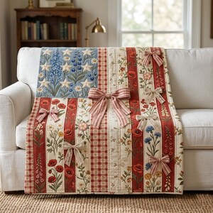 May include: A quilted throw blanket featuring a patriotic flag design with a blue field of white stars and floral motifs, alternating with vertical red and white striped panels decorated with botanical prints and several attached fabric bows.