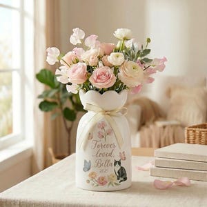 Puede incluir: Un jarrón de cerámica blanca con borde festoneado, lleno de flores rosas y blancas. El jarrón está adornado con una cinta color crema y presenta un diseño floral con mariposas y el texto "Forever loved, Bella 2025".