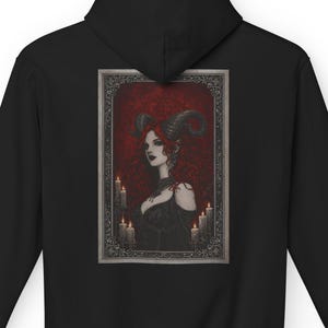 May include: Black hooded sweatshirt featuring a framed gothic illustration on the back. The artwork depicts a woman with red hair, horns, and a dark dress, surrounded by candles. The design has a dark, fantasy theme.