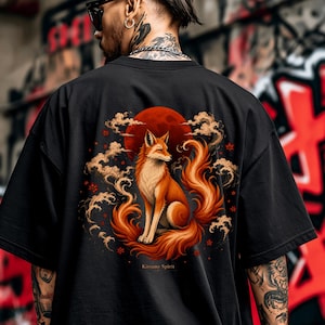 Puede incluir: Una camiseta negra con un estampado gr&aacute;fico en la espalda de un zorro naranja sentado frente a una gran luna roja, rodeado de nubes estilizadas y peque&ntilde;as flores rojas, con el texto Kitsune Spirit impreso debajo de la ilustraci&oacute;n.