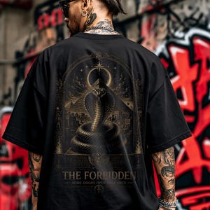 Puede incluir: Una camiseta negra de manga corta con un gran gráfico en la espalda. El diseño muestra una cobra enroscada dentro de un marco arqueado, sobre un fondo de pirámides. El texto dice THE FORBIDDEN y SOME DOORS OPEN ONLY ONCE.